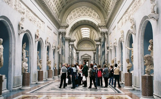 Group of visitors inside the Vatican Museums, surrounded by classical sculptures displayed in arched alcoves along an intricately decorated hall with a coffered ceiling.