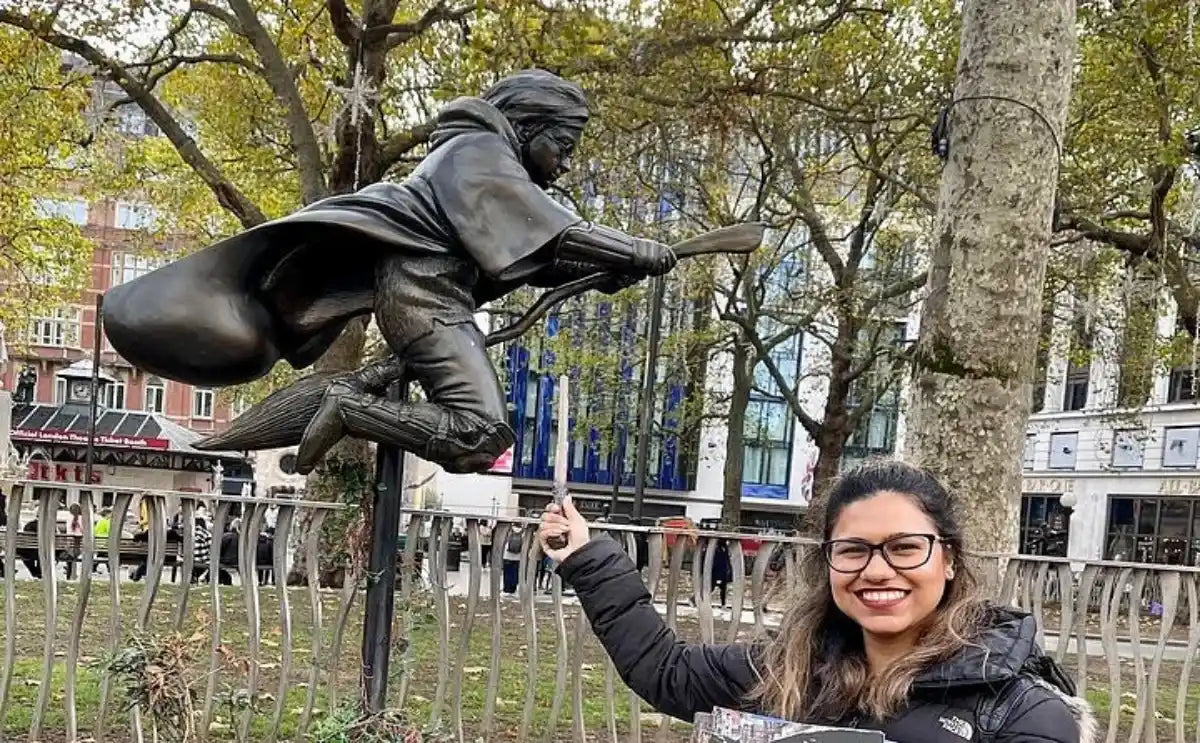 A fan smiling next to a Harry Potter-themed statue of a character on a broomstick, set in a park with trees and city buildings in the background.