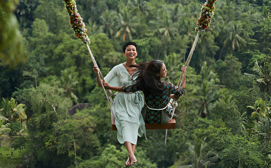 Two women on a large jungle swing in Bali, smiling as they enjoy the lush green tropical forest and serene atmosphere around them.