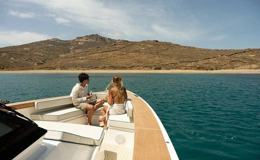A couple enjoys a relaxed moment on the deck of a luxury motor yacht, overlooking the crystal-clear waters and the serene, rugged coastline of Mykonos. The spacious seating area highlights the yacht's modern design and comfort, perfect for a private cruise experience.