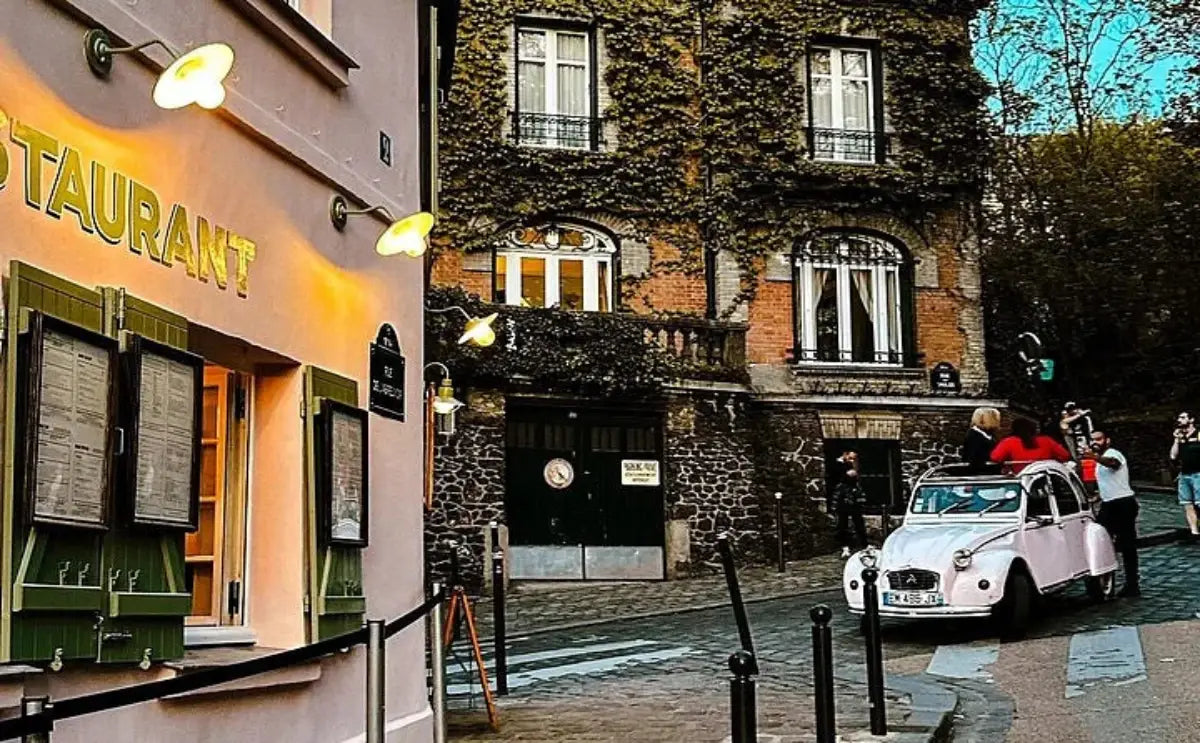 Restaurant facade in Montmartre with green shutters and vintage-style lighting, next to a cobblestone street featuring an ivy-covered building and a pink vintage car with tourists.