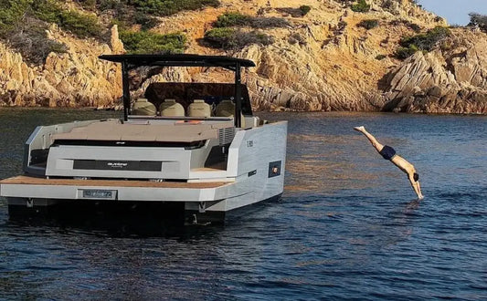 A luxurious motor yacht anchored near a rocky coastline. A guest is diving into the crystal-clear waters from the swim platform, surrounded by serene natural beauty.