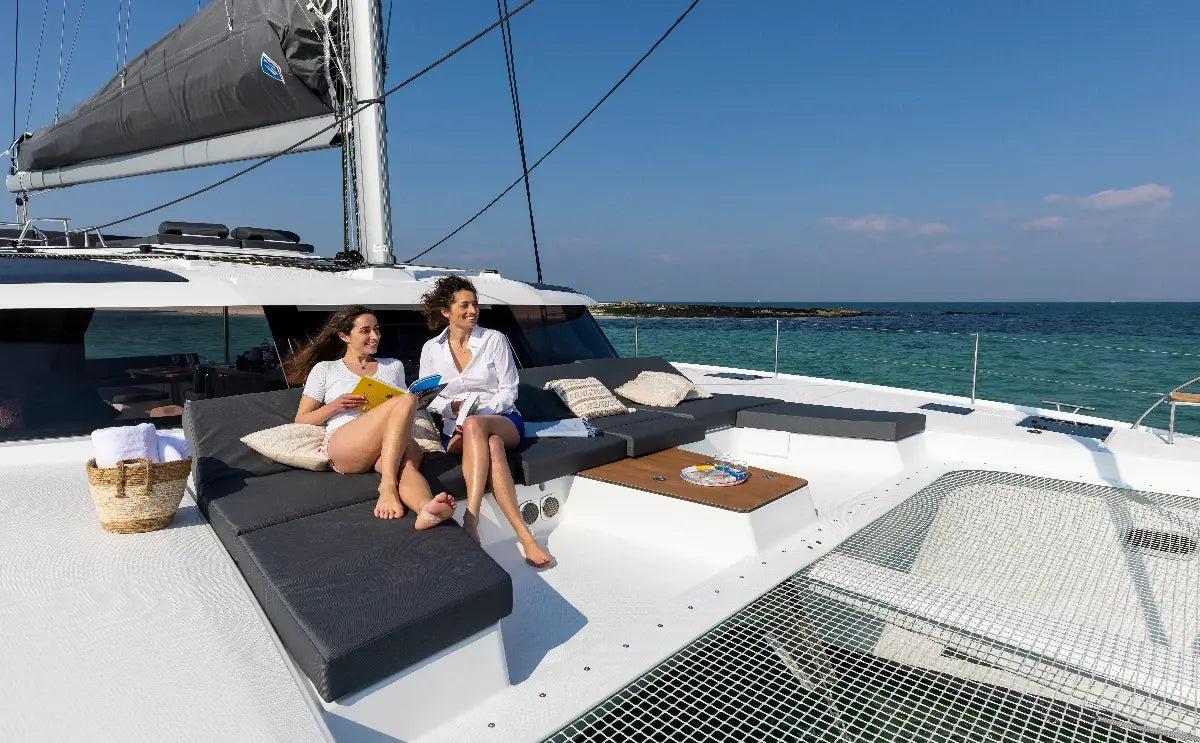 Two women relax on a spacious sailing catamaran, enjoying the sunshine and the scenic views of the turquoise waters. Luxurious cushions and refreshments enhance the leisurely atmosphere.