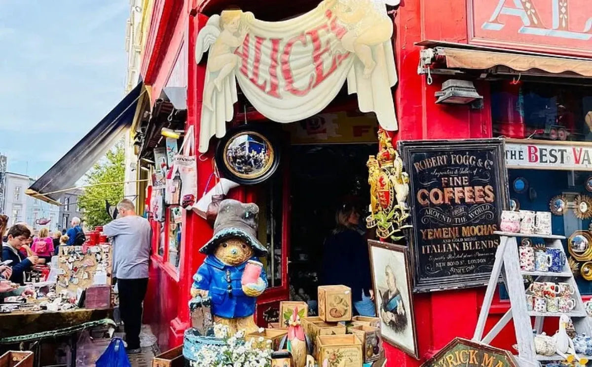 A vibrant shopfront at Portobello Market, showcasing a Paddington Bear statue, vintage signs, and an assortment of trinkets and collectibles, with shoppers browsing nearby stalls.
