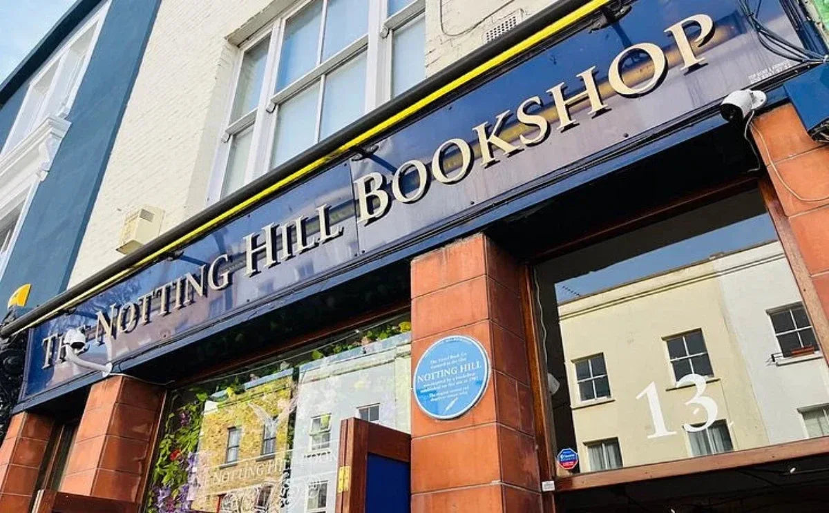 The iconic Notting Hill Bookshop facade, featuring its recognizable blue and gold sign, with a glimpse of the surrounding neighborhood reflecting in the windows.