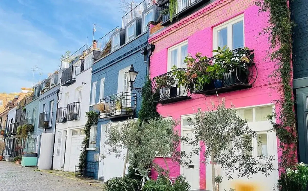 A row of colorful houses in Notting Hill, with a bright pink façade standing out among blues and whites, lined by potted plants and trees on a cobblestone street.