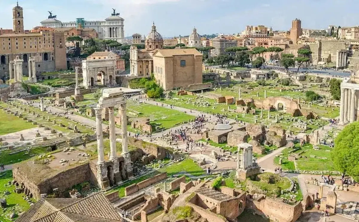 Aerial view of the Roman Forum in Rome, featuring ancient ruins such as columns, temples, and arches scattered across a grassy landscape. Visitors walk along designated paths. The Vittorio Emanuele II Monument and surrounding city buildings are visible in the background.