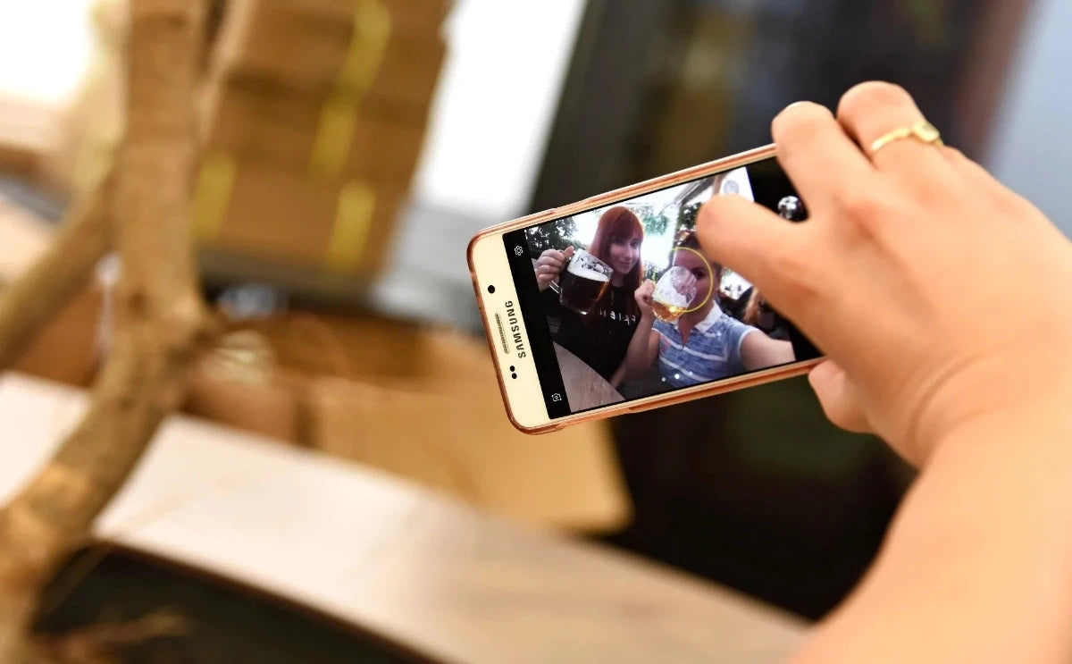 A close-up of a hand holding a smartphone, capturing a selfie of two friends enjoying glasses of Czech beer. The screen displays their cheerful expressions and raised mugs, highlighting the social and fun atmosphere of the beer tour.