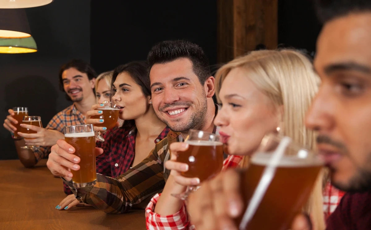 A group of people sitting at a bar, enjoying glasses of Czech beer. The man in the center is smiling directly at the camera, creating a lively and social atmosphere. The scene captures the camaraderie and enjoyment typical of a beer-tasting experience.