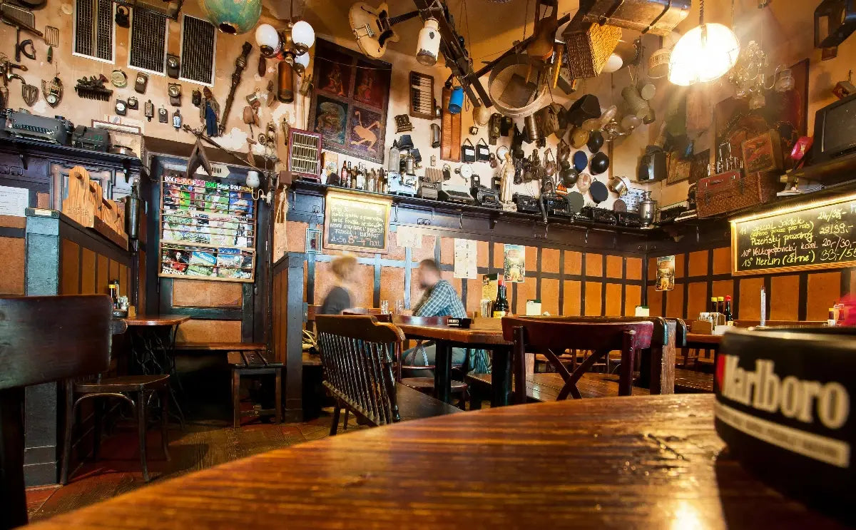 A cozy Czech pub interior featuring wooden tables and chairs, walls adorned with vintage decorations, and an eclectic mix of hanging items like guitars and kitchen utensils. A chalkboard menu is visible, and two patrons are seated at a table, adding to the relaxed atmosphere.