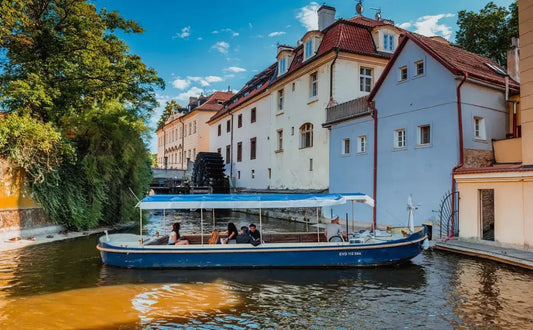 A small canal boat with a canopy carrying a group of passengers cruises through Prague's Devil's Channel, surrounded by historic buildings with red-tiled roofs and lush greenery. A watermill can be seen in the background, adding charm to the serene atmosphere.