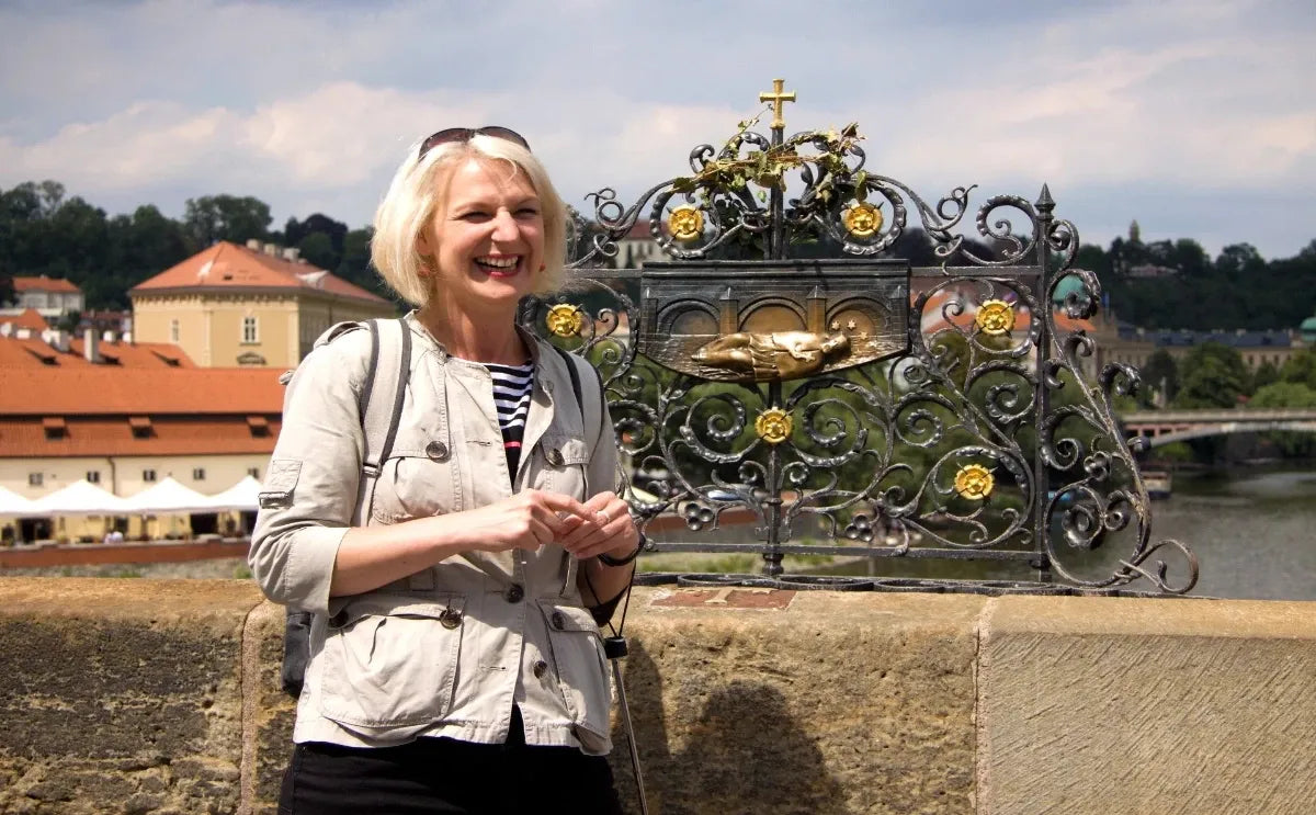 Tour guide standing on Charles Bridge in Prague, smiling beside the ornate wrought-iron railing featuring a golden relief of St. John of Nepomuk. In the background, red-roofed buildings and lush greenery are visible under a partly cloudy sky.