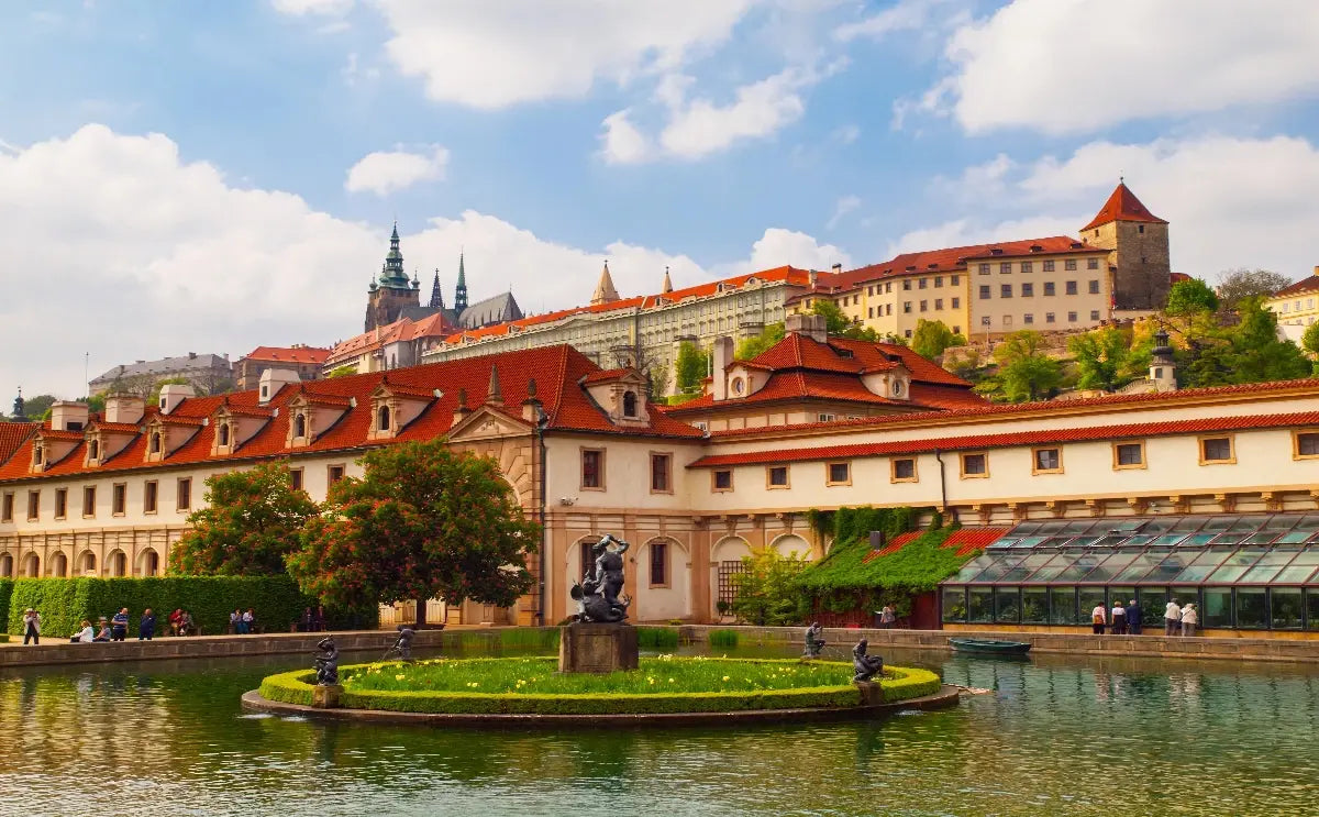 View of the Prague Castle complex with its Gothic spires and surrounding historic buildings, as seen from the serene Wallenstein Garden. The garden features a central fountain surrounded by sculptures, lush greenery, and red-roofed Baroque architecture under a partly cloudy sky.