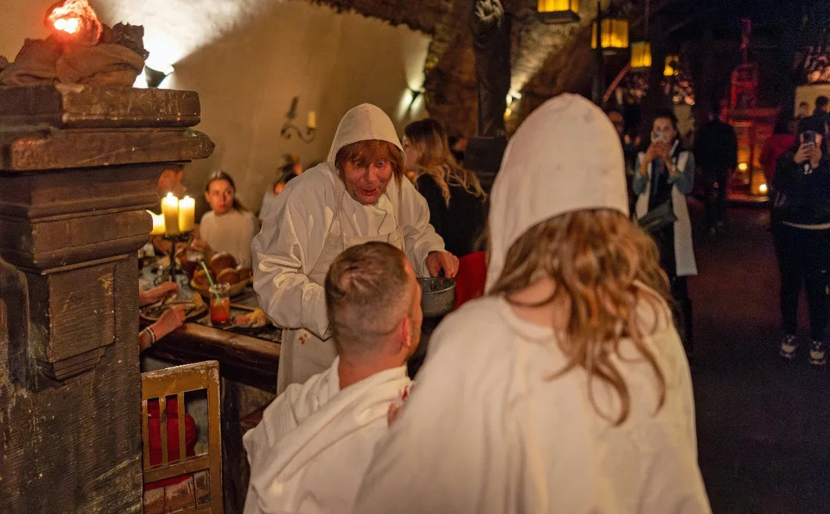 A medieval dinner scene at the Dětenice Medieval Tavern with a costumed server in a white hooded robe interacting theatrically with seated guests. The dimly lit environment includes a stone interior, candlelit tables, and other diners enjoying the immersive experience.