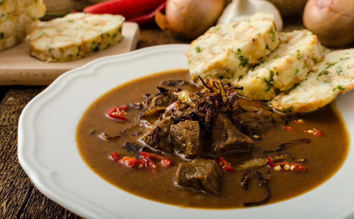A plate of traditional Czech goulash served with tender beef chunks in a rich, spiced gravy, garnished with crispy fried onions and chili slices. Accompanied by slices of fluffy bread dumplings, perfect for soaking up the flavorful sauce.