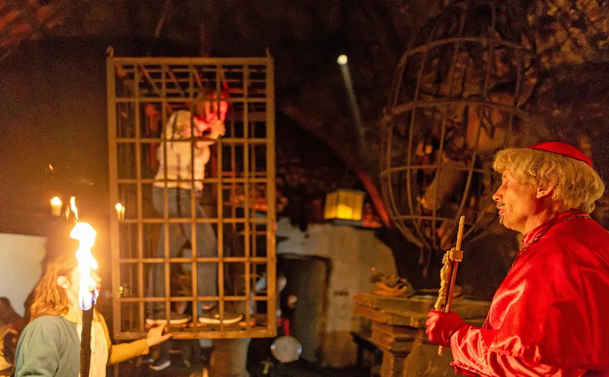 A medieval-themed scene at the Dětenice Medieval Tavern featuring a performer dressed as a cardinal holding a staff, a person in a cage as part of the theatrical display, and flaming torches illuminating the dimly lit atmosphere.