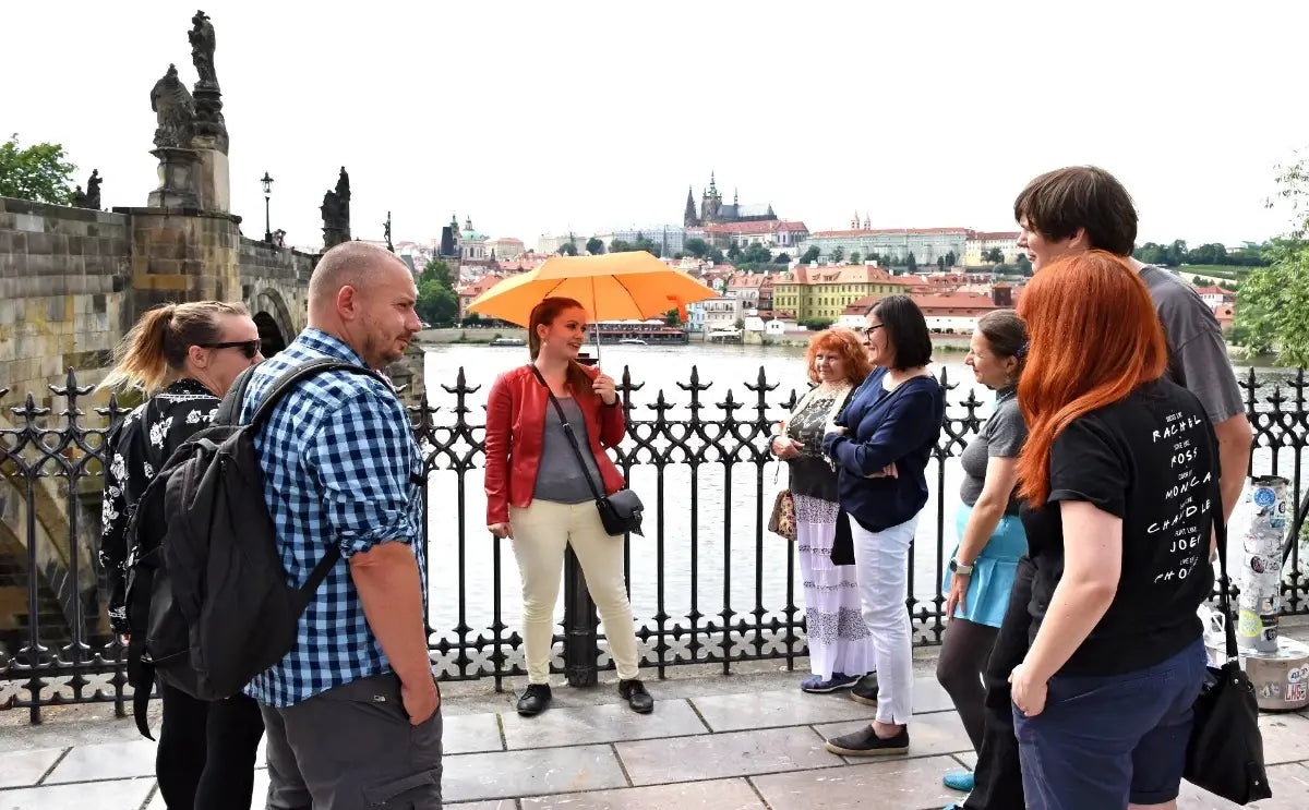 Tour guide holding an orange umbrella, speaking to a group of tourists on Charles Bridge in Prague. The group stands near an ornate black railing, with statues lining the bridge and Prague Castle visible in the background, framed by historic buildings and the Vltava River.