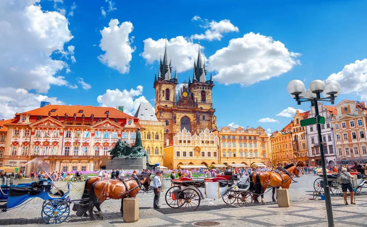 Prague's Old Town Square on a sunny day, featuring horse-drawn carriages and vibrant Renaissance-style buildings with intricate facades. The iconic Church of Our Lady Before Týn towers in the background, its Gothic spires piercing the blue sky dotted with fluffy clouds.