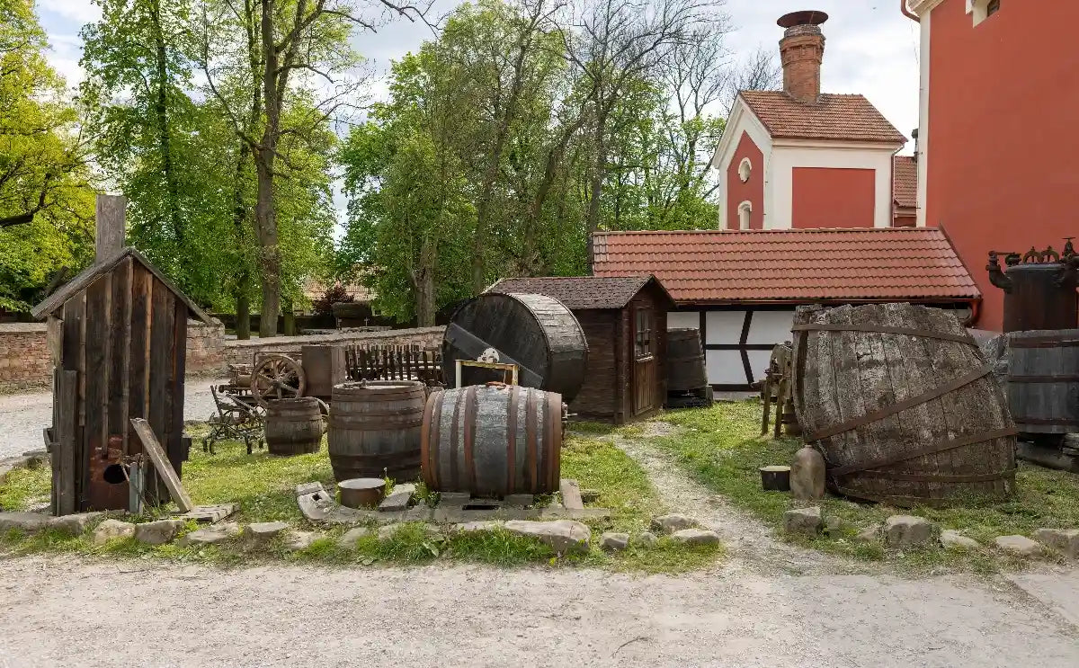 An outdoor scene at the Dětenice brewery showcasing traditional wooden barrels, brewing equipment, and rustic structures surrounded by trees, capturing the historical essence of traditional beer crafting.