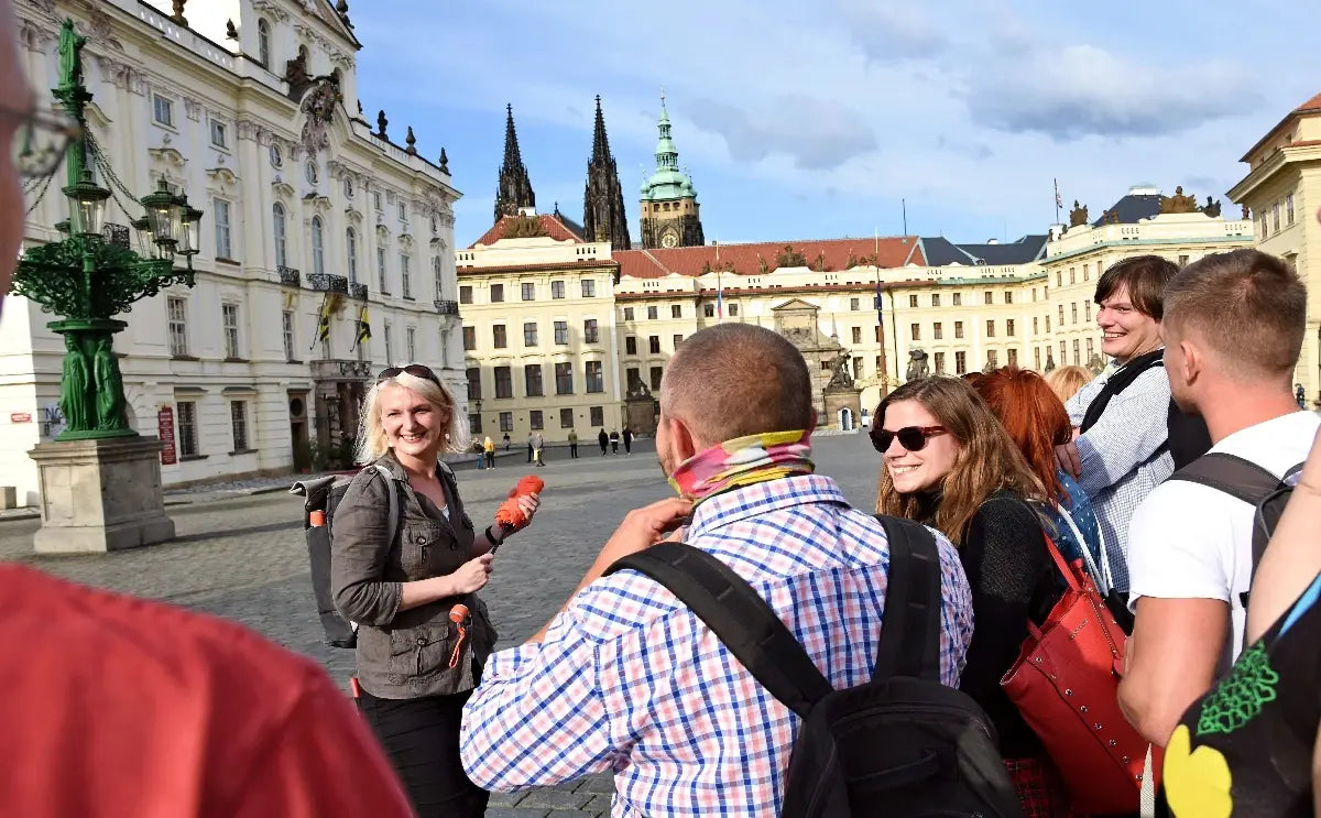 Tour guide leading a group of tourists in the courtyard of Prague Castle. The group stands in front of the grand Baroque and Gothic buildings of the castle complex, including the spires of St. Vitus Cathedral, under a partly cloudy sky.