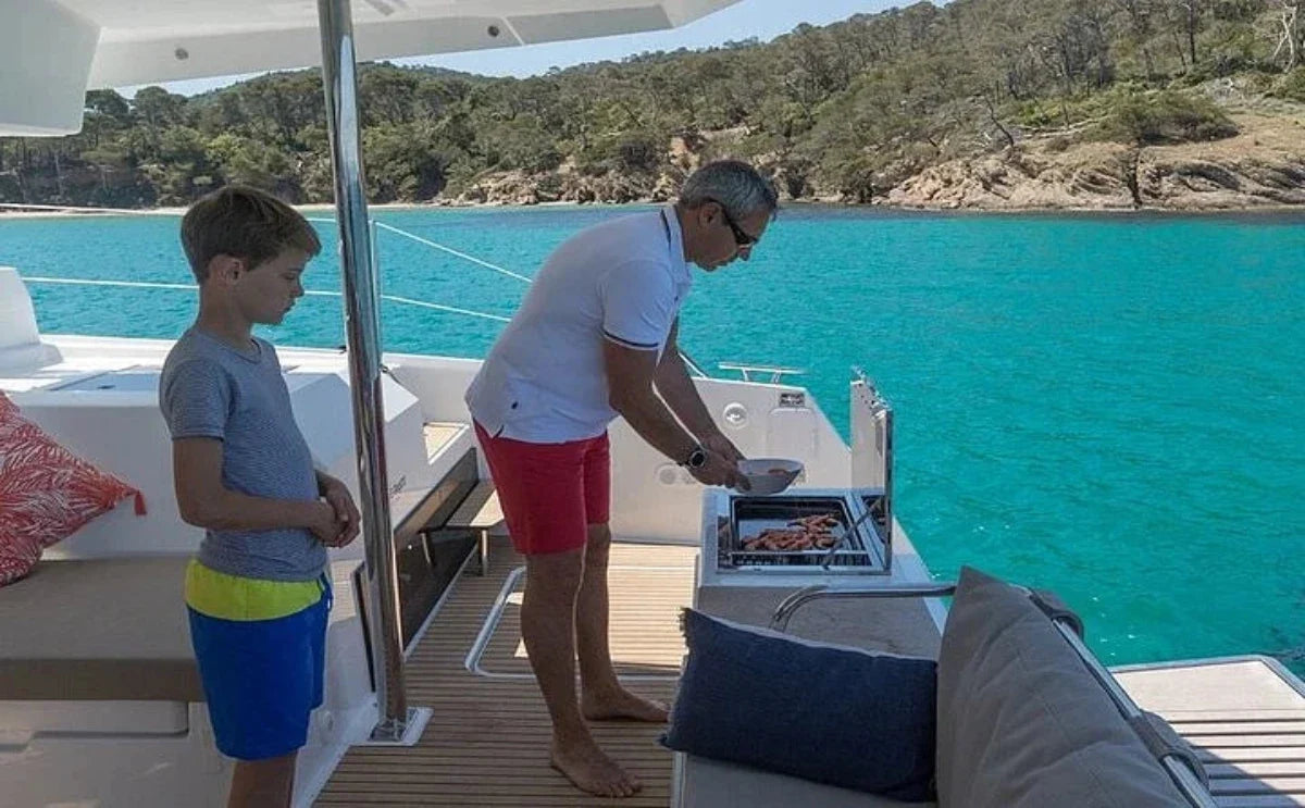 A man preparing fresh seafood on a grill aboard the "White Coral" catamaran, with a child watching, surrounded by calm turquoise waters and a scenic coastline in the background.
