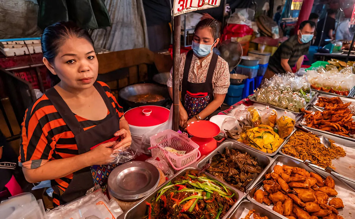 A vendor at the Maeklong Railway Market standing behind a stall filled with an array of colorful Thai street food, including fried snacks, fresh chili dishes, and packaged goods, while another vendor prepares food in the background.
