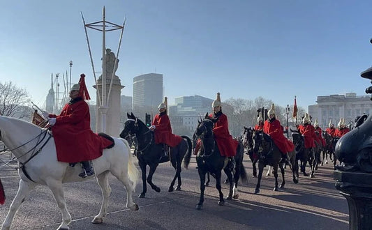A procession of mounted royal guards dressed in red coats and helmets, riding horses near a landmark in London, with city buildings in the background.