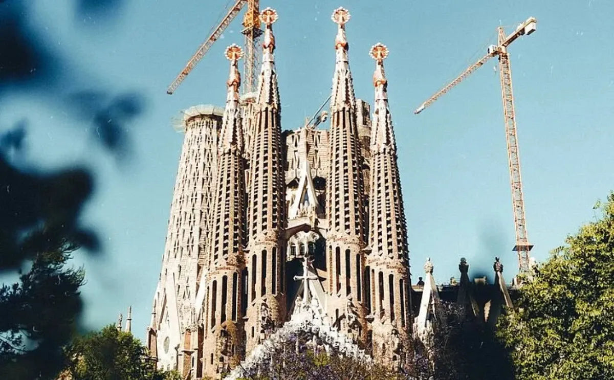 The towering spires of La Sagrada Familia dominate the skyline, with cranes visible as construction continues on Gaudí’s iconic basilica. The intricate details and unique design elements stand out against the clear blue sky, showcasing a blend of modernity and tradition.