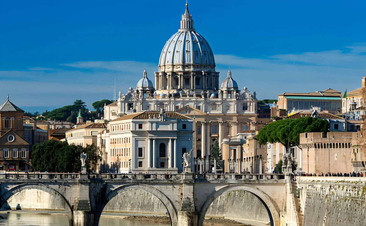 St. Peter's Basilica in Vatican City, featuring its iconic dome and ornate facade, viewed from across the Tiber River with the Ponte Sant'Angelo bridge in the foreground. The scene includes surrounding historical buildings and a clear blue sky.