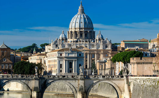 St. Peter's Basilica in Vatican City, featuring its iconic dome and ornate facade, viewed from across the Tiber River with the Ponte Sant'Angelo bridge in the foreground. The scene includes surrounding historical buildings and a clear blue sky.