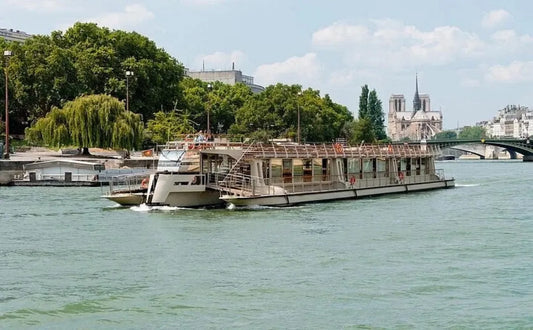 A Seine river sightseeing boat with large windows and an open deck cruising past greenery and the Notre Dame Cathedral in Paris, offering unobstructed views of iconic landmarks.
