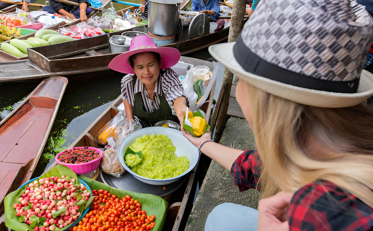 A vendor in a pink hat at the Damnoen Saduak Floating Market hands over a serving of fresh mango and sticky rice to a tourist, surrounded by colorful fruits and local delicacies in wooden boats.