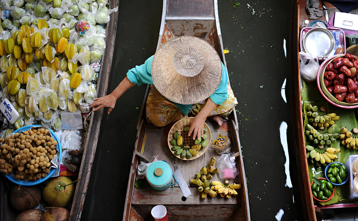 Overhead view of a vendor wearing a straw hat selling fresh tropical fruits from a wooden boat at the Damnoen Saduak Floating Market, Thailand.