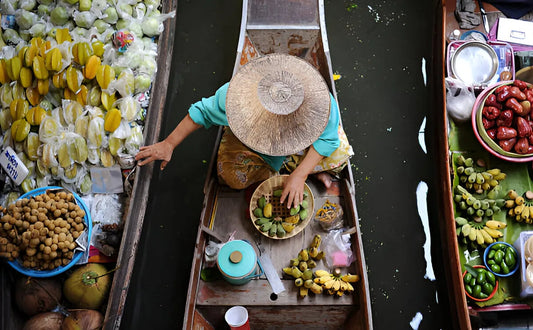 Overhead view of a vendor wearing a straw hat selling fresh tropical fruits from a wooden boat at the Damnoen Saduak Floating Market, Thailand.